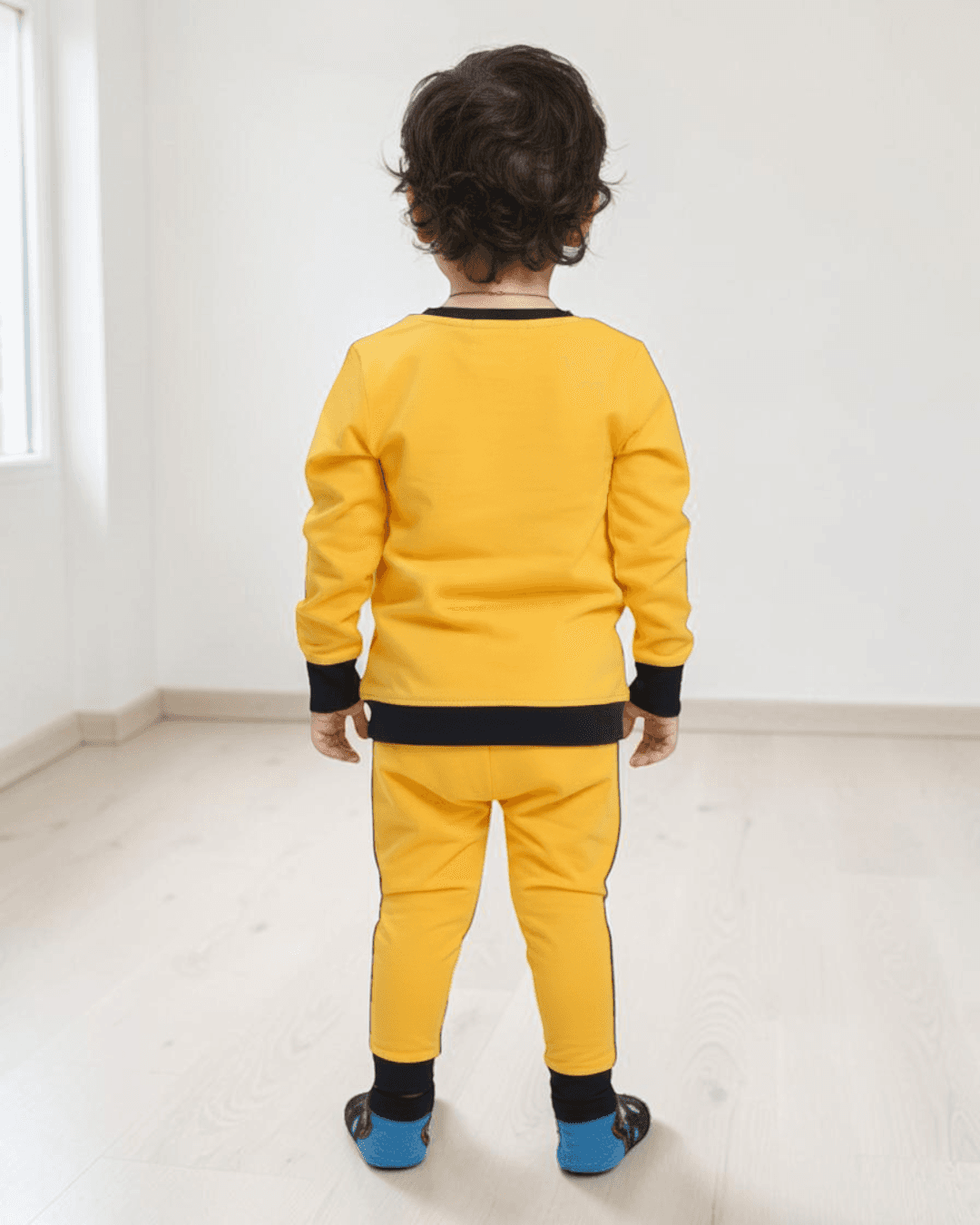 Back view of a young boy wearing yellow kids' leggings with black cuffs, paired with a matching yellow sweatshirt, standing in a bright room