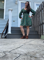 Model wearing a green floral Donna Shirred Dress outdoors. Features a smocked bodice, long sleeves, and a flared skirt, perfect for casual or dressy occasions.