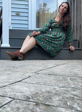Model sitting on outdoor steps in a green floral Donna Shirred Dress. This mini dress features a shirred bodice, long sleeves, and a flared skirt for a stylish look.