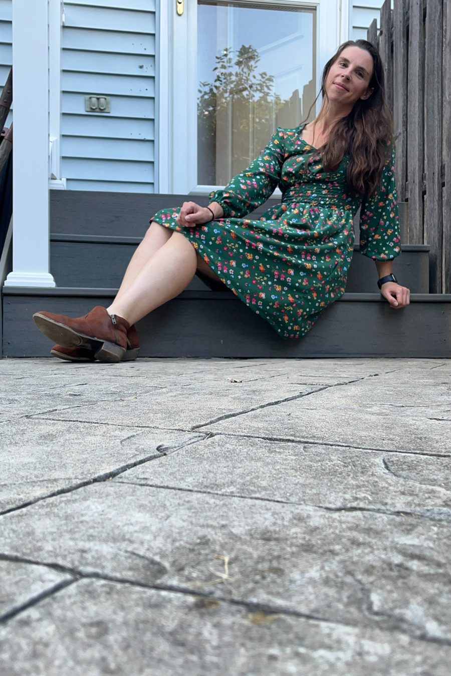 Model sitting on outdoor steps in a green floral Donna Shirred Dress. This mini dress features a shirred bodice, long sleeves, and a flared skirt for a stylish look.